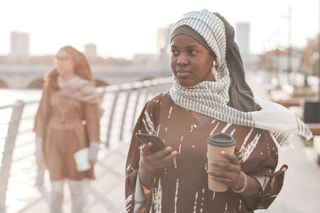 African muslim woman drinking coffee and using mobile phone during her walk in the cityの写真素材