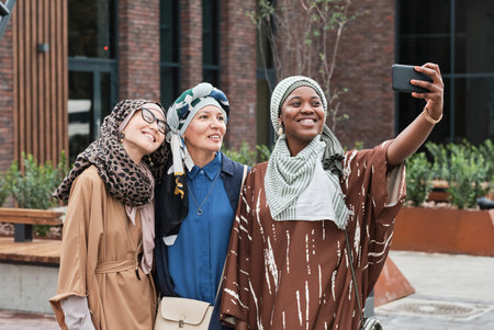 Three elegant women in muslim clothes smiling at camera of mobile phone while making selfie portrait outdoorsの写真素材
