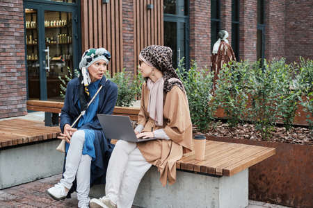Young muslim woman sitting on the bench typing on laptop and discussing something together with other woman outdoorsの写真素材