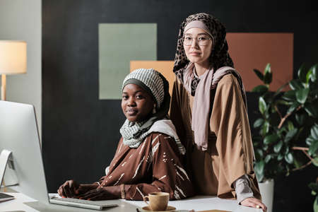 Portrait of two muslim businesswomen looking at camera while working on computer in team at officeの写真素材