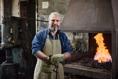 Portrait of mature carpenter in apron looking at camera standing near the furnace in the factoryの写真素材