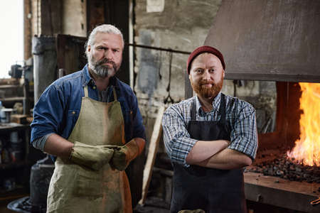 Portrait of two carpenters in aprons looking at camera while working in the workshopの写真素材