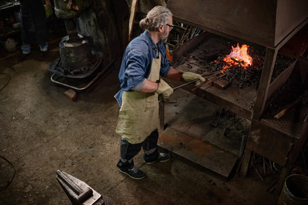 Worker putting coal into furnace for making details from iron in the workshop