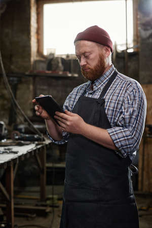 Mature engineer in apron typing on digital tablet during his work in workshopの写真素材