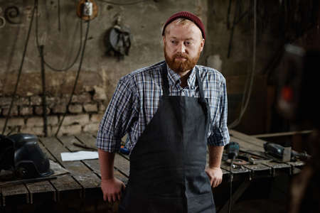 Craftsman in apron standing near the workplace with tools and resting after work in the workshopの写真素材