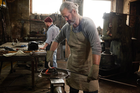 Mature bearded blacksmith making detail from iron together with colleague in the background in the blacksmith shopの写真素材