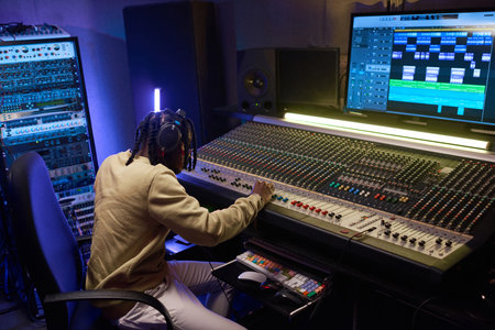 African musical producer in headphones working on sound mixing board in digital recording studio