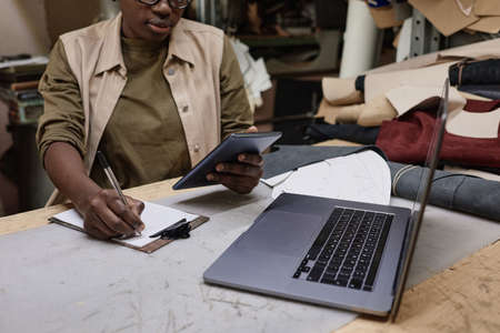 African tailor sitting at her workplace with laptop using tablet pc and making notes in document,...