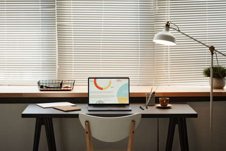 Image of workplace with laptop with graphs on the screen standing in front of the window with...
