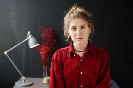Portrait of pretty young woman in red shirt looking at camera standing at modern room with workplace...