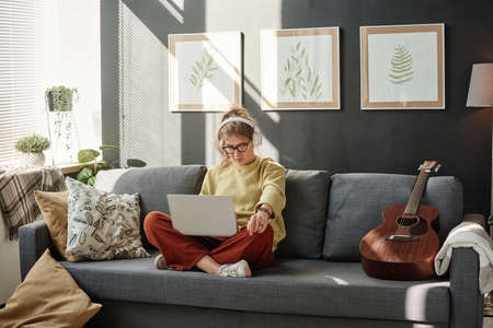 Young woman in headphones watching something on laptop during her leisure time on the sofa in the...