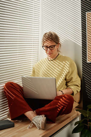 Young woman in eyeglasses sitting on windowsill and communicating online using her laptopの写真素材