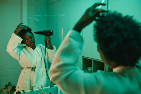 African young woman in bathrobe looking at mirror and drying her hair with hair drier in the bathroomの写真素材