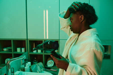 African woman in white bathrobe making hairstyle with hair dryer after bath in the morning