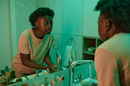 African young woman standing in the bathroom and looking at the mirror, she washing her face with waterの写真素材