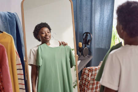 African young woman standing in front of the mirror and trying on the new dress while standing in the roomの写真素材