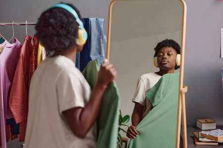 African woman in wireless headphones trying on the new dress while standing in front of the mirror in the roomの写真素材