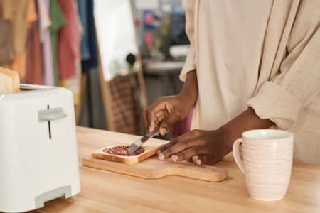 Close-up of African woman applying jam on bread while cooking at the kitchen table at homeの写真素材