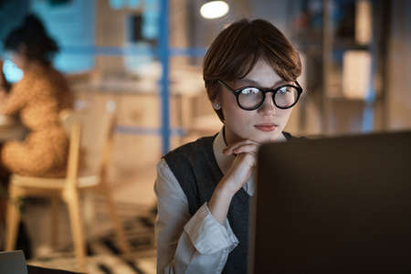 Young businesswoman in eyeglasses sitting at the table in front of the screen of laptop and working at office till late nightの写真素材