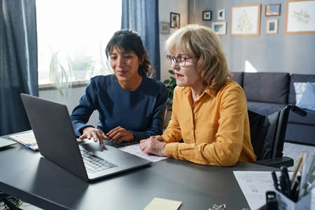 Young volunteer showing to senior woman how to use computer while they sitting at the table in the living roomの写真素材