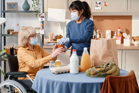 Woman in protective mask and gloves delivering food to home for senior woman who using wheelchair
