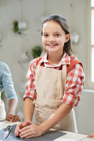 Portrait of happy little girl in apron smiling at camera while cooking at the tableの写真素材