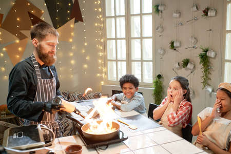 Chef in apron cooking food on the fire and showing masterclass to children in the kitchen