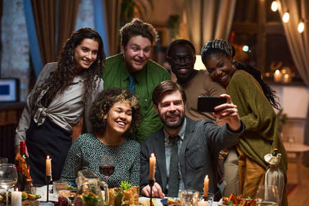 Group of happy friends smiling at camera while making selfie portrait on mobile phone at dining...