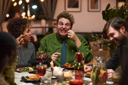 Young man joking with his friends while they sitting at the table during dinner partyの写真素材