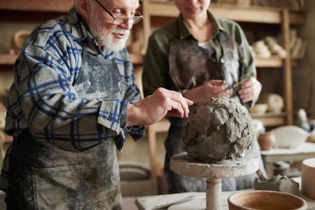 Senior potter in eyeglasses using stick to make form from piece of clay, he teaching his student of...