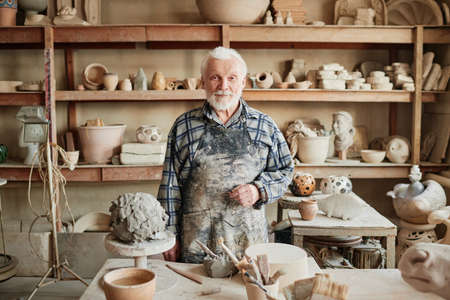 Portrait of senior potter with white beard looking at camera while making sculptures from clay in the workshopの写真素材