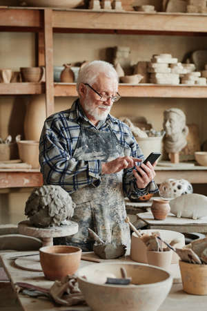 Professional senior potter taking orders by mobile phone while working with clay sculptures in...
