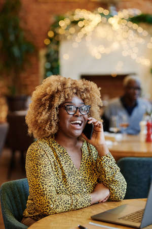 Happy African businesswoman in eyeglasses laughing while having a conversation on mobile phone sitting at the table in cafeの写真素材