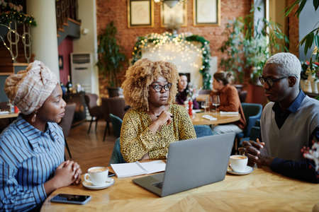 Group of African business people sitting at the table with laptop drinking coffee and discussing during meeting in cafeの写真素材