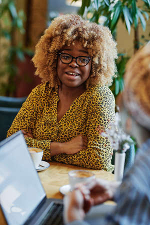 African businesswoman with curly hair talking to her colleague during coffee break at the table in cafeの写真素材