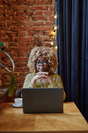 African businesswoman with curly hair sitting at the table in front of the laptop in cafe and...