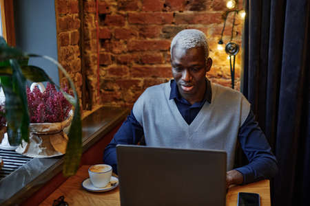 African young man working online on laptop and drinking coffee while sitting at the table in cafe