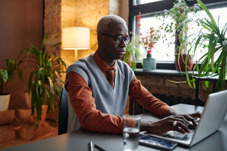 Serious African businessman in eyeglasses sitting at the table and typing on laptop working at...