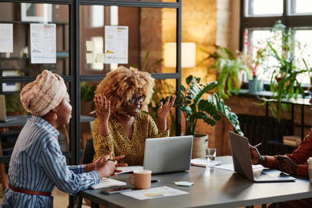 African businesswoman with curly hair sitting at the table with laptop and talking to her colleagues...