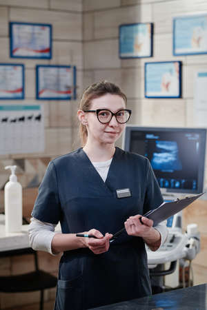 Portrait of young vet nurse in uniform holding medical card and looking at camera while working in vet clinicの写真素材