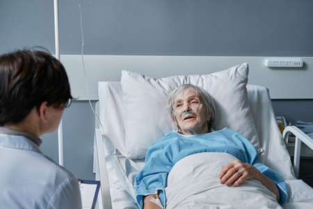 Rear view of young woman visiting her senior mother while she lying on bed at hospital ward