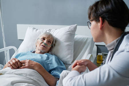 Senior woman lying on hospital bed and talking to the nurse about her disease at wardの写真素材
