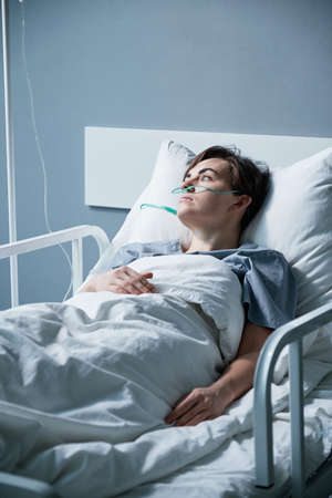 Young woman being treated at hospital, she lying on the bed with oxygen tube and looking away with pensive sightの写真素材