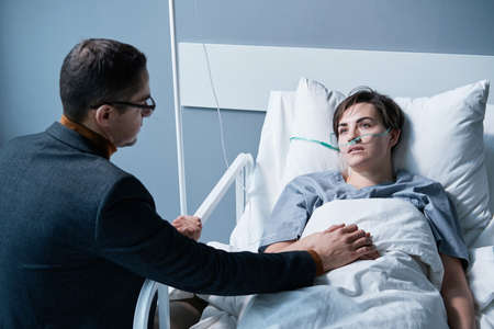 Rear view of young man supporting the sick young woman while she lying on bed at hospital wardの写真素材