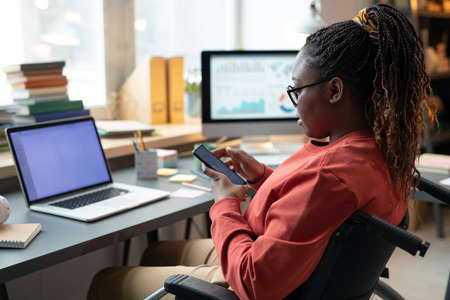 African woman with disability reading a message on mobile phone while sitting on wheelchair at the table with laptopの写真素材