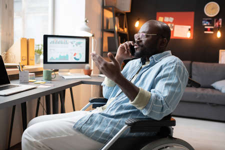 African man with disability sitting on wheelchair and talking on mobile phone, he working at home at the table with laptopの写真素材