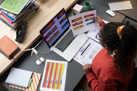 High angle view of African businesswoman sitting at the table in front of the laptop and examining charts and graphs at officeの写真素材
