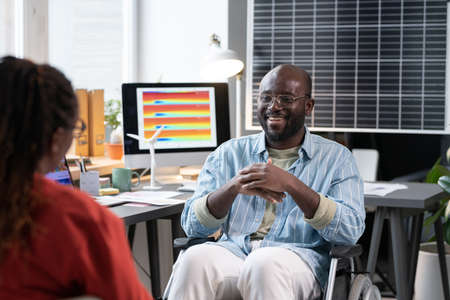 African man sitting on wheelchair and talking to his colleague during break at officeの写真素材