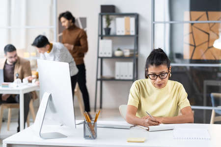 Young businesswoman in eyeglasses sitting at her workplace with computer and making notes in notebook during her work at officeの写真素材