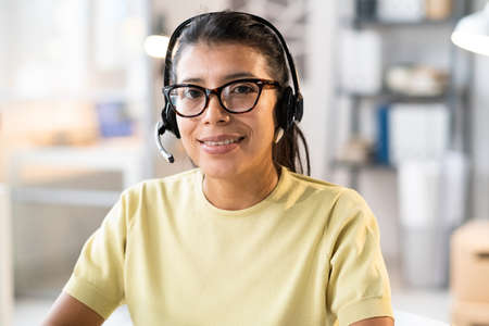 Portrait of call centre operator in headphones smiling at camera while working with clients at...